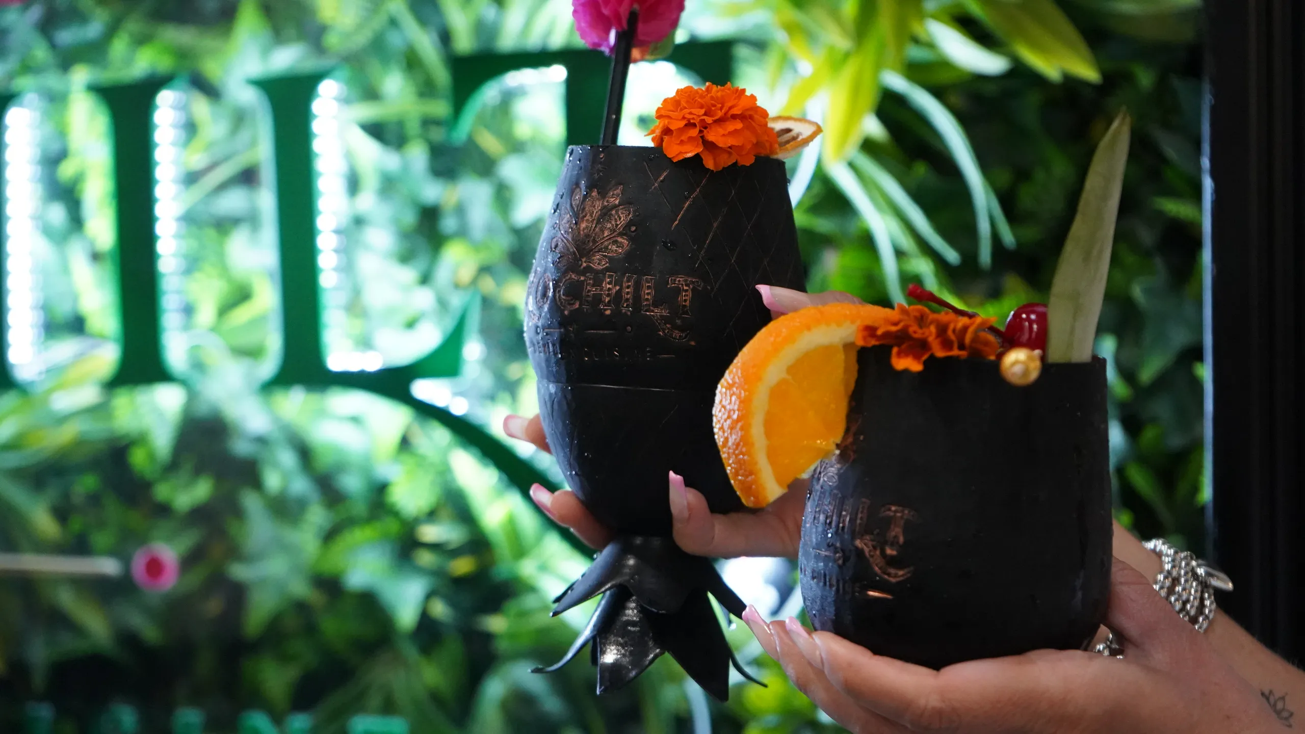 Two engraved Xochitl cocktail cups garnished with orange wheels and marigolds, raised in front of the restaurant's living-plant wall and glowing sign