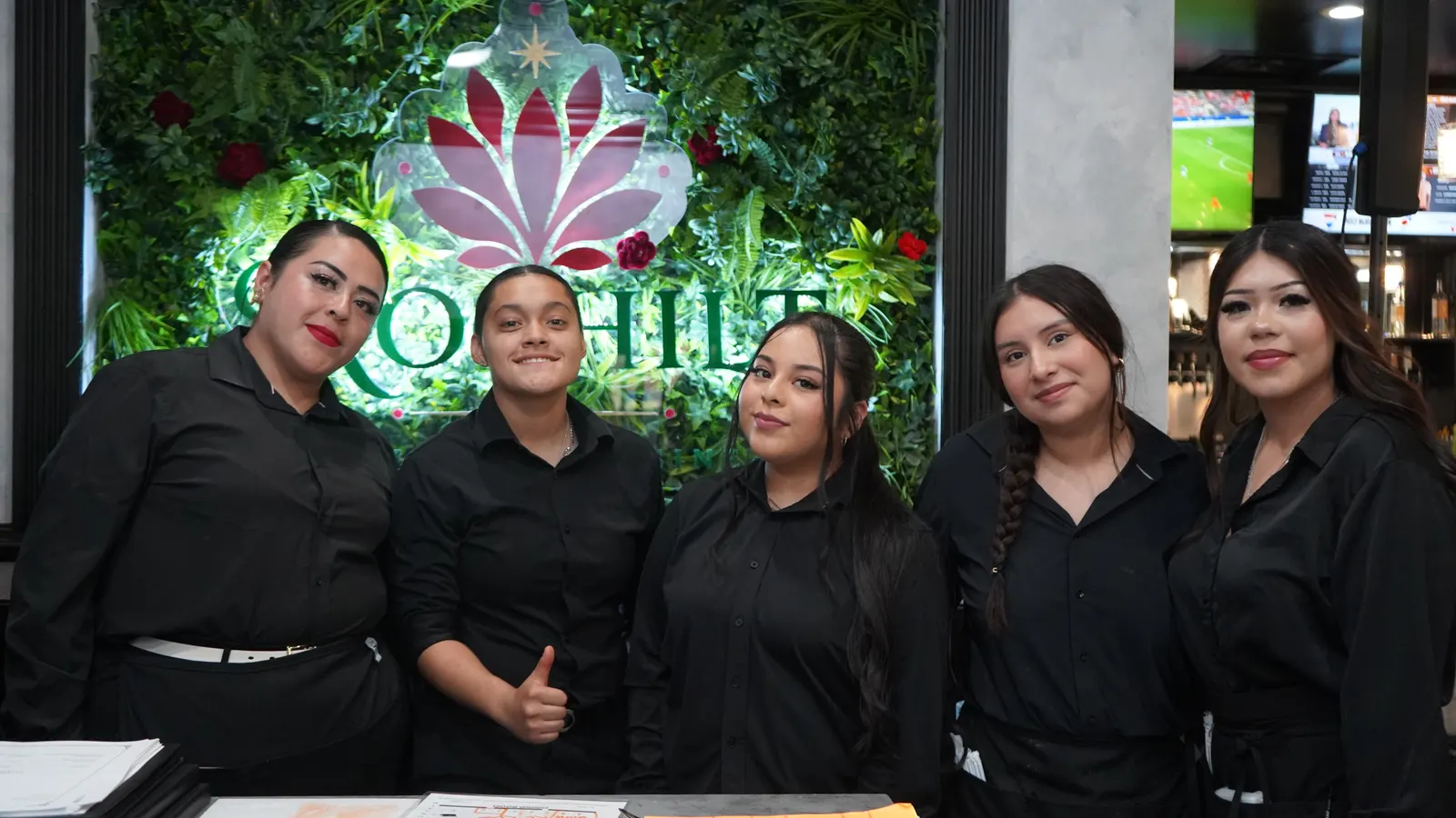Five smiling Xochitl team members in black at the host stand, in front of the living-plant wall and flower logo